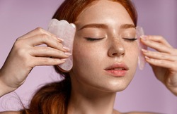 Close up portrait of beautiful redhead girl, ginger female massaging face with jade scraper, close eyes, doing tcm cosmetology procedure, purple background