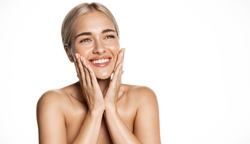 Spa and cosmetology. Young blond woman smiling happy, looking up, holding hands on face, apply moisturizing cream, face lotion or toner, white background
