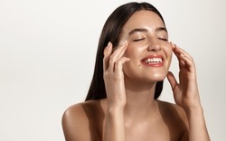 Smiling woman with clean glowing facial skin washing her face, rubbing in cream skincare product for anti-aging, lifting spa effect, standing over white background