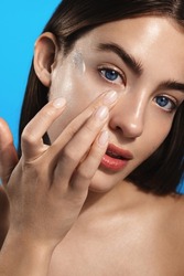 Close up of young woman with clear glowing skin, looks in mirror, applies facial lotion, cream or acne treatment on face, stands with bare shoulders on blue background