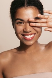 Vertical of dark skinned model shows her glowing, clear natural facial skin, covers her eye with one hand and smiles at camera, brown background