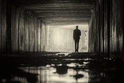 Silhouette of a person with a lantern in a square dark concrete drainage tunnel with light in the end.