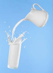 Pouring milk from jug into glass cup with splashing on blue background