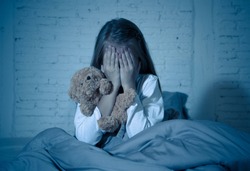 Scared little girl sitting in bed covering her face with hands holding her teddy in fear afraid of monsters in darkness in bedroom in Child nightmares imagination and psychological distress concept.