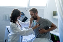Young sick man patient with Oxygen Mask while female doctor listens his chest with stethoscope in hospital emergency room. In Smoking and respiratory diseases and anti tobacco advertising campaign.