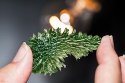 Rare wrinkled green moldavite mineral in human hand with orange light on black background. Collector holding meteoric glass gem from Czechia at incandescent lamp with beautiful bokeh. Selective focus.
