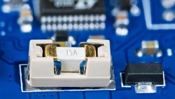 Small electrical fuse in white holder and black linear fixed voltage regulator on blue PCB. Printed circuit board detail with surface mount technology of electronic components and bokeh in background.