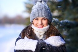 Portrait of happy positive girl, young beautiful attractive pretty woman is freezing walking outdoors in winter snowy park at cold frosty day in hat, scarf, gloves in snow, smiling, looking at camera