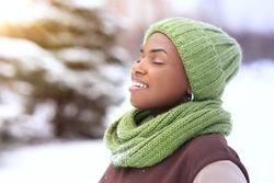 Portrait of beautiful attractive happy girl young black African Afro American woman is enjoying good cold frosty sunny day at winter snow park with eyes closed, breathing deeply deep fresh air smiling