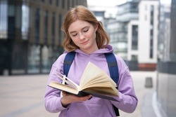 Young beautiful blonde woman, college or university academic student girl or school pupil is studying, preparing to lesson, reading book or textbook outdoors at campus with backpack 