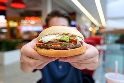 Close up photo of tasty delicious big burger or hamburger, sandwich with beef in man hands at food court in shopping mall. Guy is holding out fast junk food in hands. 