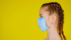 Teen girl in a medical mask in profile looks away on yellow background with copy space. Epidemic precautions. Acute Respiratory Virus Infection. Illness cold flu. Protective mask.