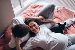 Soft focus. Pregnant woman, man and dog lying on a bed near the window. Attractive couple look to each other and smiling.  Natural light, terracotta bed linen. close up shot.