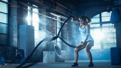 Athletic Female in a Gym Exercises with Battle Ropes During Her Cross Fitness Workout/ High-Intensity Interval Training. She's Muscular and Sweaty, Gym is in Deserted Factory. Cold Ambient.