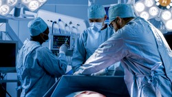 Diverse Team of Professional Surgeons Performing Invasive Surgery on a Patient in the Hospital Operating Room. Nurse Hands Out Instruments to surgeon,  Anesthesiologist Monitors Vitals. Cold and Blue.
