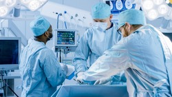 Diverse Team of Professional Surgeons Performing Invasive Surgery on a Patient in the Hospital Operating Room. Nurse Hands Out Instruments to surgeon,  Anesthesiologist Monitors Vitals.