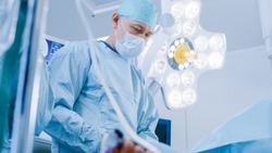 Low Angle Shot in Operating Room, Assistant Passes Instrument to a Chief Surgeon During Invasive Surgery. Professional Doctors Performing Surgery in Modern Hospital.