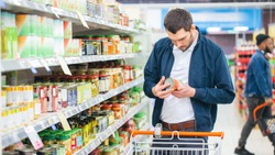 At the Supermarket: Handsome Man Uses Smartphone and Looks at Nutritional Value of the Canned Goods. He's Standing with Shopping Cart in Canned Goods Section.