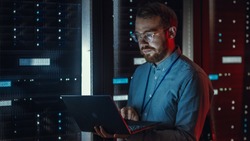 Bearded IT Specialist in Glasses is Working on Laptop in Data Center while Standing Near Server Rack. Running Diagnostics, Doing Maintenance Work. Emergency Red Light from Side Illuminating Specialist