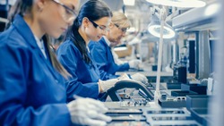 Female Electronics Factory Workers in Blue Work Coat and Protective Glasses Assembling Printed Circuit Boards for Smartphones with Tweezers. High Tech Factory with Employees.