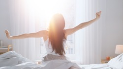 Beautiful Brunette is Waking up in the Morning, Stretches in the Bed, Sun Shines on Her From the Big Window. Happy Young Girl Greets New Day with Warm Sunlight Flare.