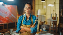 Professional Young Female Artist Dirty with Paint, Wearing Apron, Arms Crossed while Holding Brushes, Looks at the Camera with a Smile. Authentic Creative Studio with Large Canvas. Face Portrait