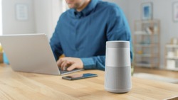 Close Up Shot of a Modern Silver Bluetooth Speaker Standing on a Table at Home. Man in the Background Sits at the Table and Works on His Laptop. Smartphone Lies on a Table Next to Him.
