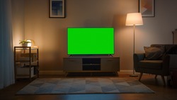 Shot of a TV with Horizontal Green Screen Mock Up. Cozy Evening Living Room with a Chair and Lamps Turned On at Home.