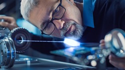 Close-up Portrait of Focused Middle Aged Engineer in Glasses Working with High Precision Laser Equipment, Using Lenses and Optics for Accuracy Electronics. Testing Superconductor Material