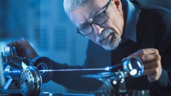 Close-up Portrait of Focused Middle Aged Engineer in Glasses Working with High Precision Laser Equipment, Using Lenses and Testing Optics for Accuracy Required Electronics