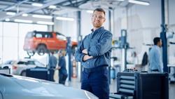 Handsome Car Mechanic is Posing in a Car Service. He Wears a Jeans Shirt and Safety Glasses. His Arms are Crossed. Specialist Looks at a Camera and Smiles.