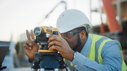 On the Commercial / Industrial Building Construction Site: Professional Engineer Surveyor Takes Measures with Theodolite, Worker Uses Laptop. In the Background Skyscraper Formwork Frames and Crane
