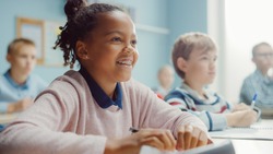 In Elementary School Class: Portrait of a Brilliant Black Girl with Braces Smiles, Writes in Exercise Notebook. Junior Classroom with Diverse Group of Children Learning New Stuff