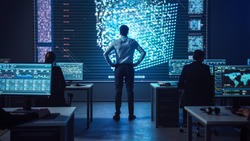 Project manager Stands Next to Big Screen with Neural Network. Team of Professional Computer Data Science Engineers Work on Desktops in a Dark System Control and Monitoring Telecommunications Office.