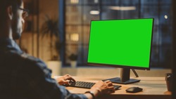 Over the Shoulder: Creative Young Man Sitting at His Desk Using Desktop Computer with Mock-up Green Screen. Evening in the Stylish Office Studio with City Window View