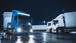 Blue Semi-Truck with Cargo Trailer Drives Off From Overnight Parking Space where Other Trucks are Standing. Long Haul Truck Leaves Parking Lot, Transporting Cargo / Goods Across Continent. Rainy Night