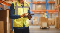 Male Worker Wearing Hard Hat Checks Products Stock and Inventory with Digital Tablet Standing in Retail Warehouse full of Shelves with Goods. Distribution, Logistics. Close-up Shot.