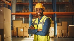 Handsome and Happy Professional Worker Wearing Safety Vest and Hard Hat Smiling with Crossed Arms on Camera. In the Background Big Warehouse with Shelves full of Delivery Goods. Medium Portrait
