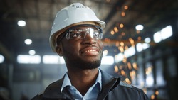 Happy Professional Heavy Industry Engineer Worker Wearing Uniform, Glasses and Hard Hat in a Steel Factory. Smiling African American Industrial Specialist Standing in a Metal Construction Manufacture.