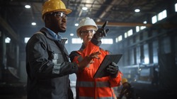 Two Heavy Industry Engineers Stand in Steel Metal Manufacturing Factory, Use Digital Tablet Computer and Have a Discussion. Black African American Industrial Specialist Talk to Female Technician.