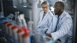 Bright Medical Science Laboratory with Diverse Team of Research Scientists Working. Young Female Microbiologist Talks to a Male Biochemist about New Test Results. High-Tech Equipment.
