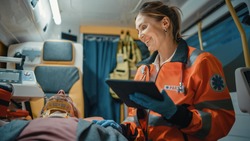 Female EMS Professional Paramedic Using Tablet Computer to Fill a Questionnaire for the Injured Patient on the Way to Hospital. Emergency Care Assistant Comforting the Patient in an Ambulance.