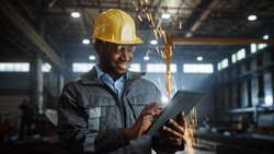 Professional Heavy Industry Engineer Worker Wearing Safety Uniform and Hard Hat Uses Tablet Computer. Smiling African American Industrial Specialist Standing in a Metal Construction Manufacture.