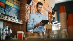 Handsome Caucasian Coffee Shop Owner is Working on Tablet Computer and Checking Inventory in a Cozy Loft-Style Cafe. Successful Restaurant Manager Standing Happy Behind Counter.