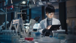 Medical Development Laboratory: East Asian Scientist Uses Digital Tablet Computer, Uses Microscope, Conducting Experiment. Pharmaceutical Lab for Research Medicine, Biotechnology. Evening Work