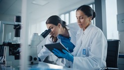 Modern Medical Research Laboratory: Two Female Scientists Working Together Using Microscope, Analyzing Samples, Talking. Advanced Scientific Pharmaceutical Lab for Medicine, Biotechnology Development