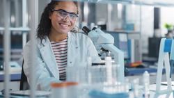 Medical Science Laboratory: Portrait of Black Scientist Using Microscope for Analysis of Test Sample, Smileing on Camera. Ambitious Young Biotechnology Specialist, working with Advanced Equipment
