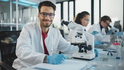 Medical Science Laboratory: Handsome Latin Scientist Analysing Samples and Uses Microscope, Looking at Camera and Smiling Charmingly. Young Biotechnology Specialist, Using Advanced Equipment.