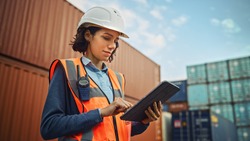 Smiling Portrait of a Beautiful Latin Female Industrial Engineer in White Hard Hat, High-Visibility Vest Working on Tablet Computer. Inspector or Safety Supervisor in Container Terminal.