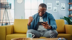 Happy Black African American Man Playing Video Game on Smartphone App and Win, Showing YES Gesture. He is Sitting on a Sofa and Resting in Living Room, Having Fun Over the Internet.